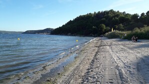 A sandy beach with footprints leading to the water, a forested hill in the background, and a clear sky. - Charming house in medieval village with pond view terrace, private parking (Miramas)