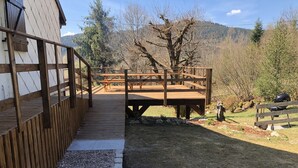 Una terraza de madera que conduce a un área de picnic con mesas, rodeada por un paisaje natural con árboles y colinas.
