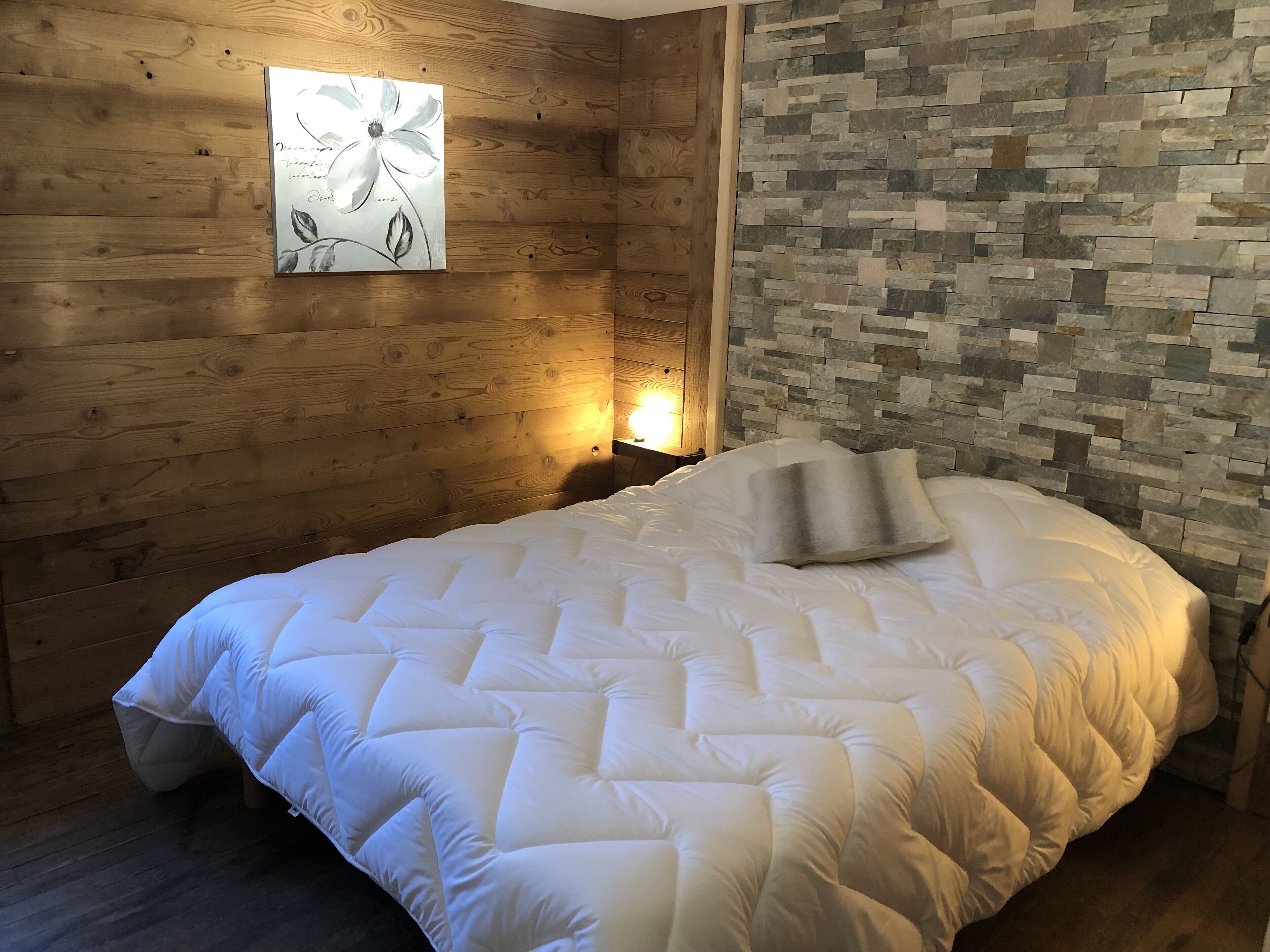 A bedroom with a wooden wall, a stone wall, a bed with white bedding, and a wall-mounted artwork.