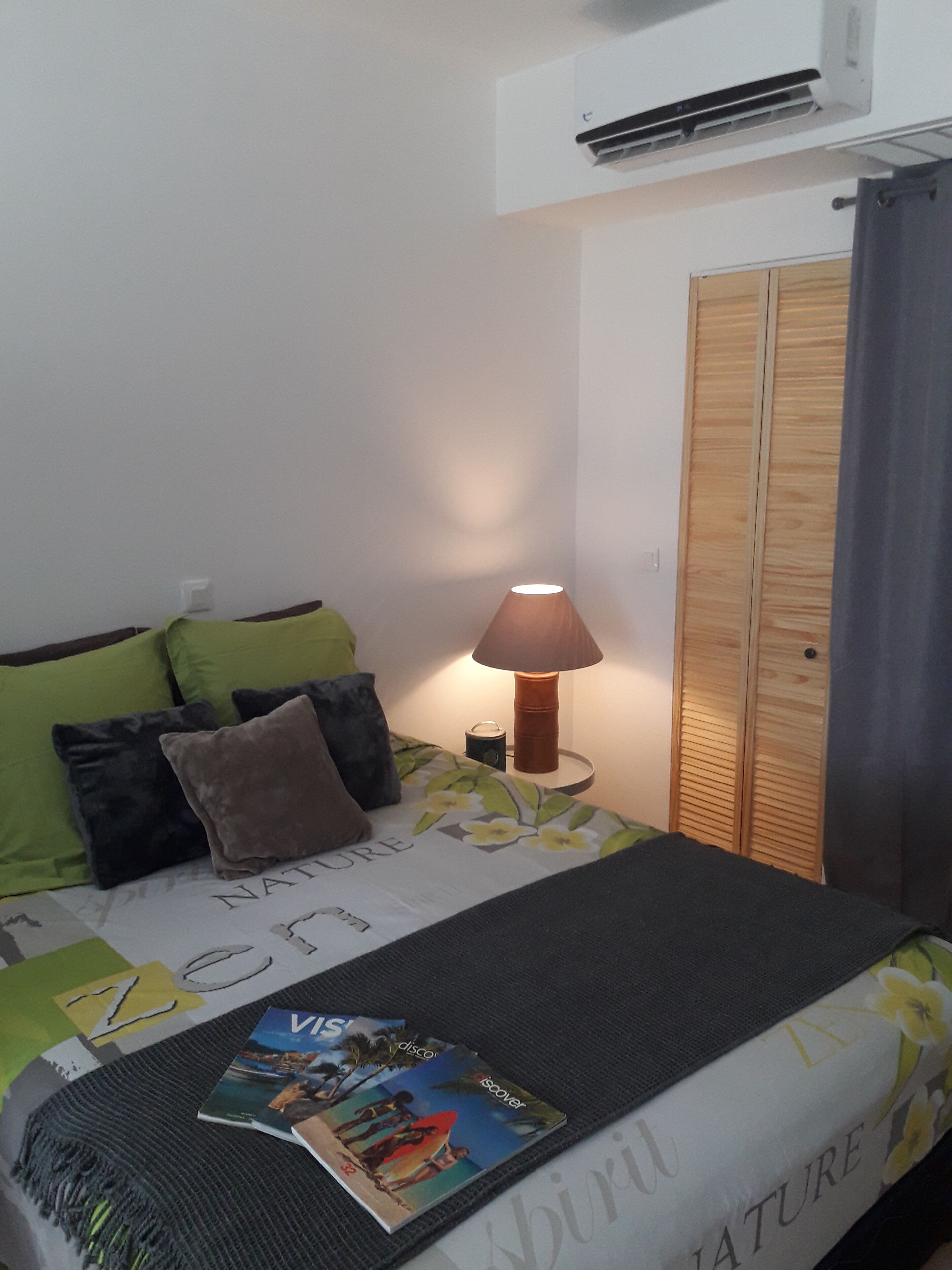 A bedroom with a bed, pillows, a bedside lamp, a magazine, a wardrobe, and an air conditioning unit.