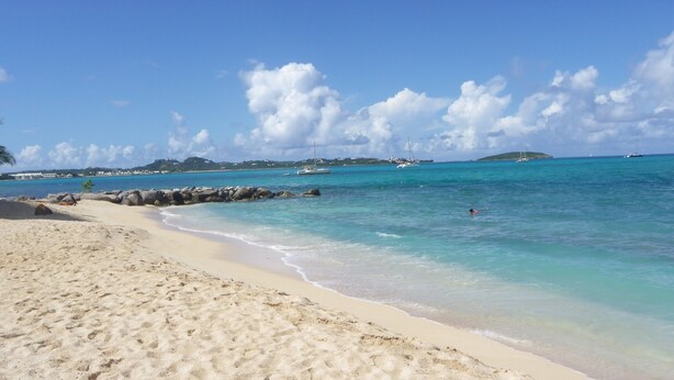 A sandy beach with clear turquoise water, a rocky breakwater, and distant islands.