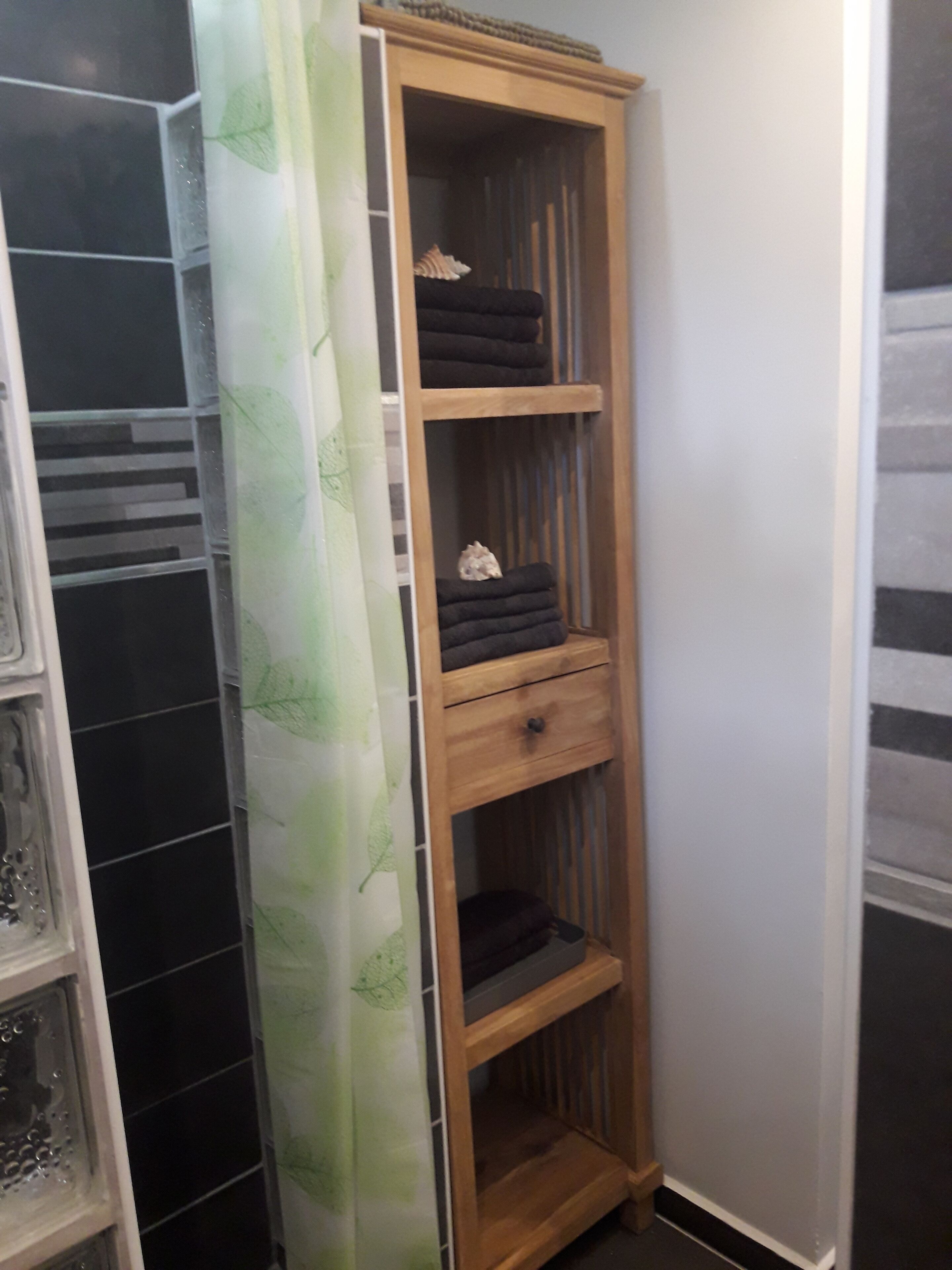 Une armoire en bois, dotée de plusieurs étagères et d’un tiroir, est placée dans un coin, contre un mur recouvert de carreaux noirs et devant un rideau à motifs de feuilles vertes.