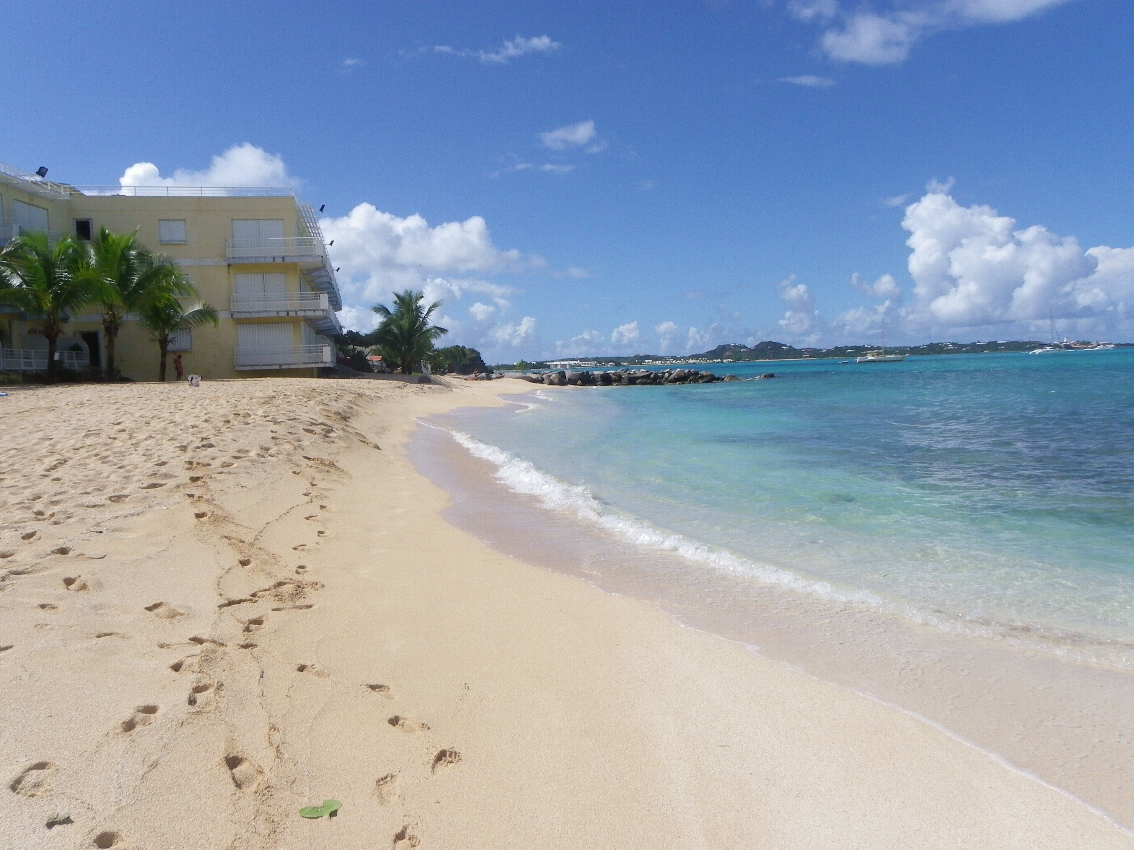 A sandy beach with footprints, a building with balconies, palm trees, and a clear blue sky.