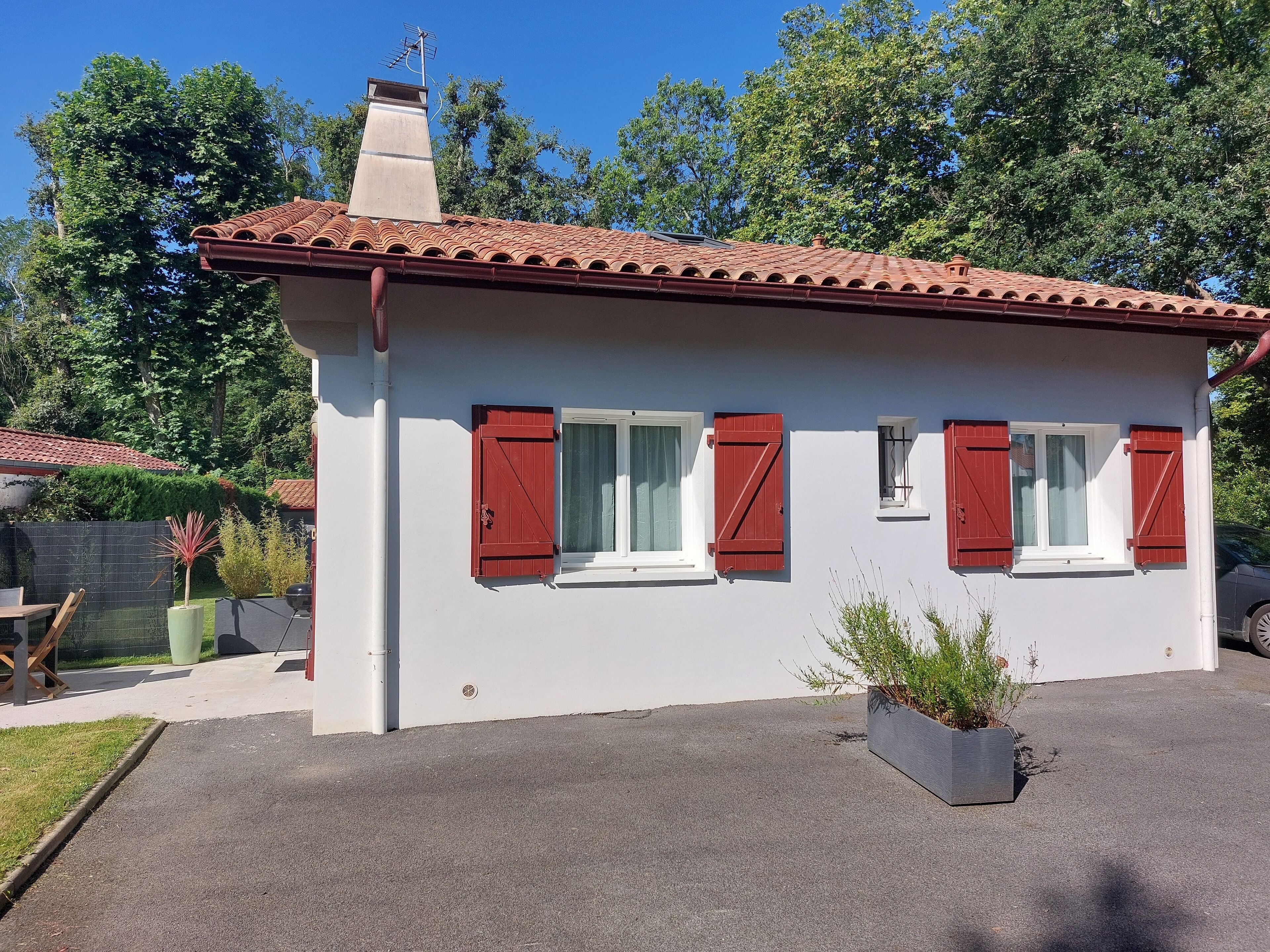 Une petite maison à un étage, avec un toit recouvert de tuiles rouges, des murs blancs et des volets rouges. Une cheminée se dresse sur le toit, et la maison est entourée d’une clôture et de verdure.