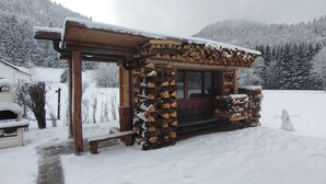 Une cabane en bois, dont le toit est recouvert de neige, avec un banc et une pile de bois de chauffage à l’extérieur.
