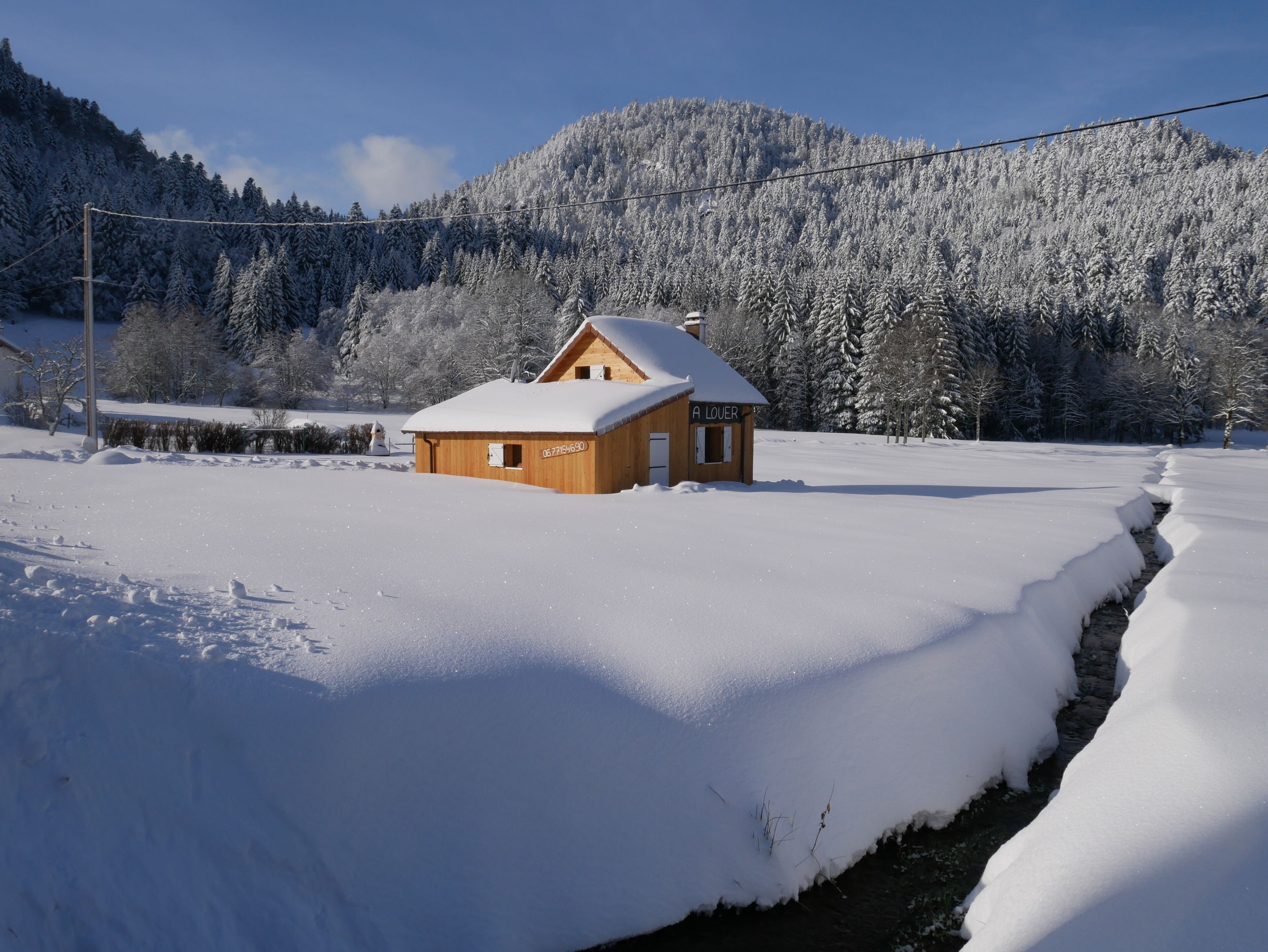 Une cabane en bois dont le toit est recouvert de neige, entourée d’un paysage enneigé et avec une montagne en arrière-plan.
