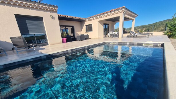 Une piscine aux eaux cristallines, bordée d’une terrasse carrelée, un patio couvert et un bâtiment à la façade beige et au toit recouvert de tuiles.
