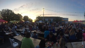 Un evento al aire libre concurrido, con mesas y sillas, carpas y un atardecer de fondo.