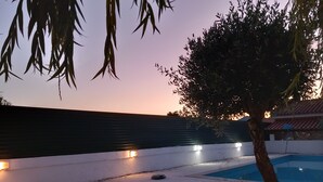 Zona de piscina con suelo de baldosas, un árbol y un edificio al fondo.