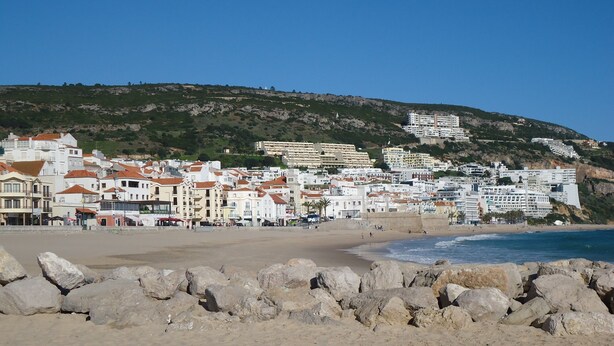 Un pueblo costero con edificios a lo largo de la playa, primer plano rocoso y una colina al fondo.
