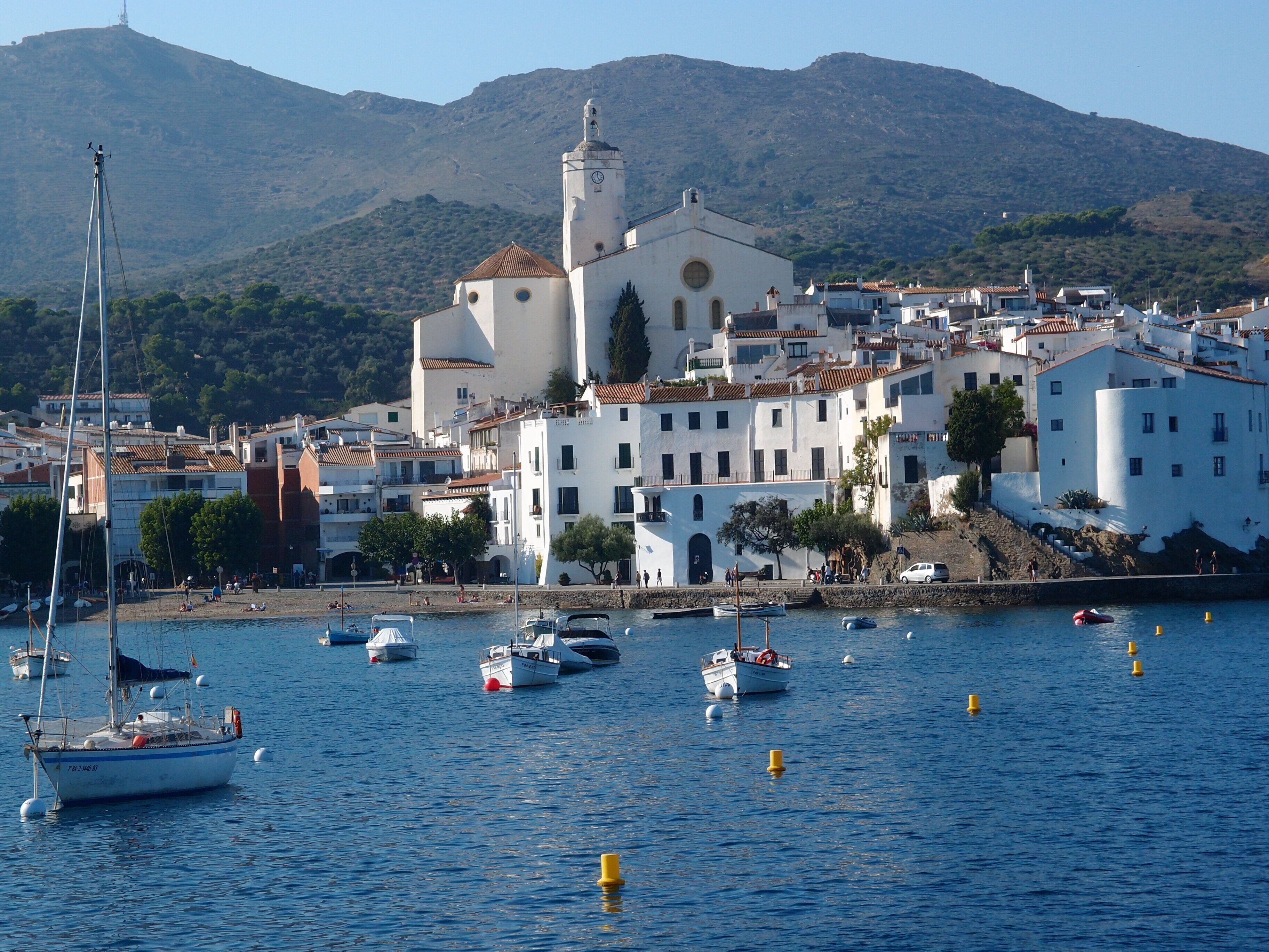 A coastal town with a prominent church, boats in the harbor, and a mountainous backdrop.