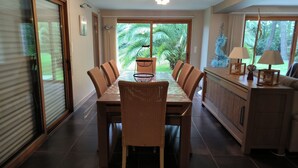 Ein Essbereich mit einem langen Tisch und Stühlen, ein Küchenbereich mit einem Schrank und einer Lampe sowie Blick ins Grüne durch die Fenster.