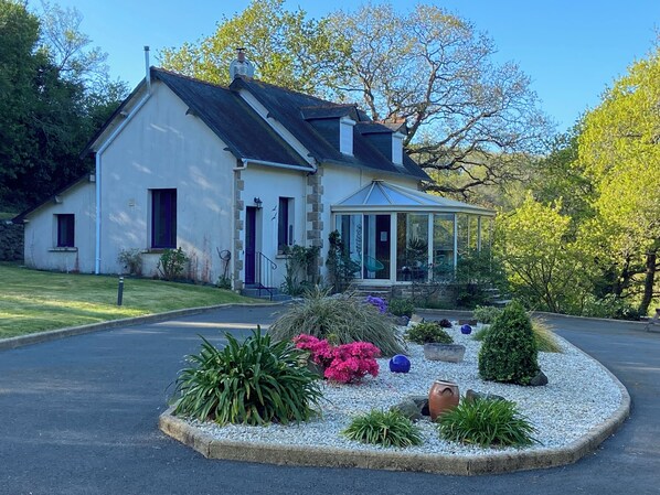 Una casa a due piani con veranda, circondata da un giardino ghiaioso con diverse piante e un piccolo stagno.