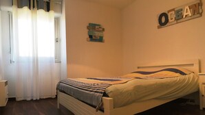 A bedroom with a bed, a nightstand, a wall-mounted shelf, and a window with curtains. - Apartment F3 1st line Carnon East, feet in the water (Mauguio)