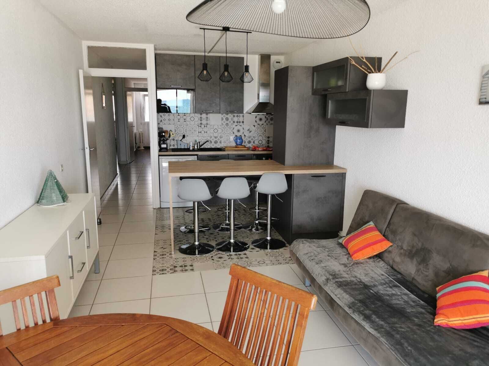 Een moderne keuken met een centraal eiland, barkrukken, een bank met kleurrijke kussens, en een eethoek met een houten tafel en stoelen.