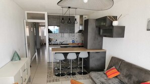A modern kitchen with a central island, bar stools, a sofa with colorful cushions, and a dining area with a wooden table and chairs. - Apartment F3 1st line Carnon East, feet in the water (Mauguio)