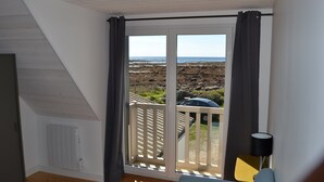 Une pièce donnant sur une plage rocheuse, avec un radiateur blanc, un rideau gris, une chaise jaune et une lampe noire.
