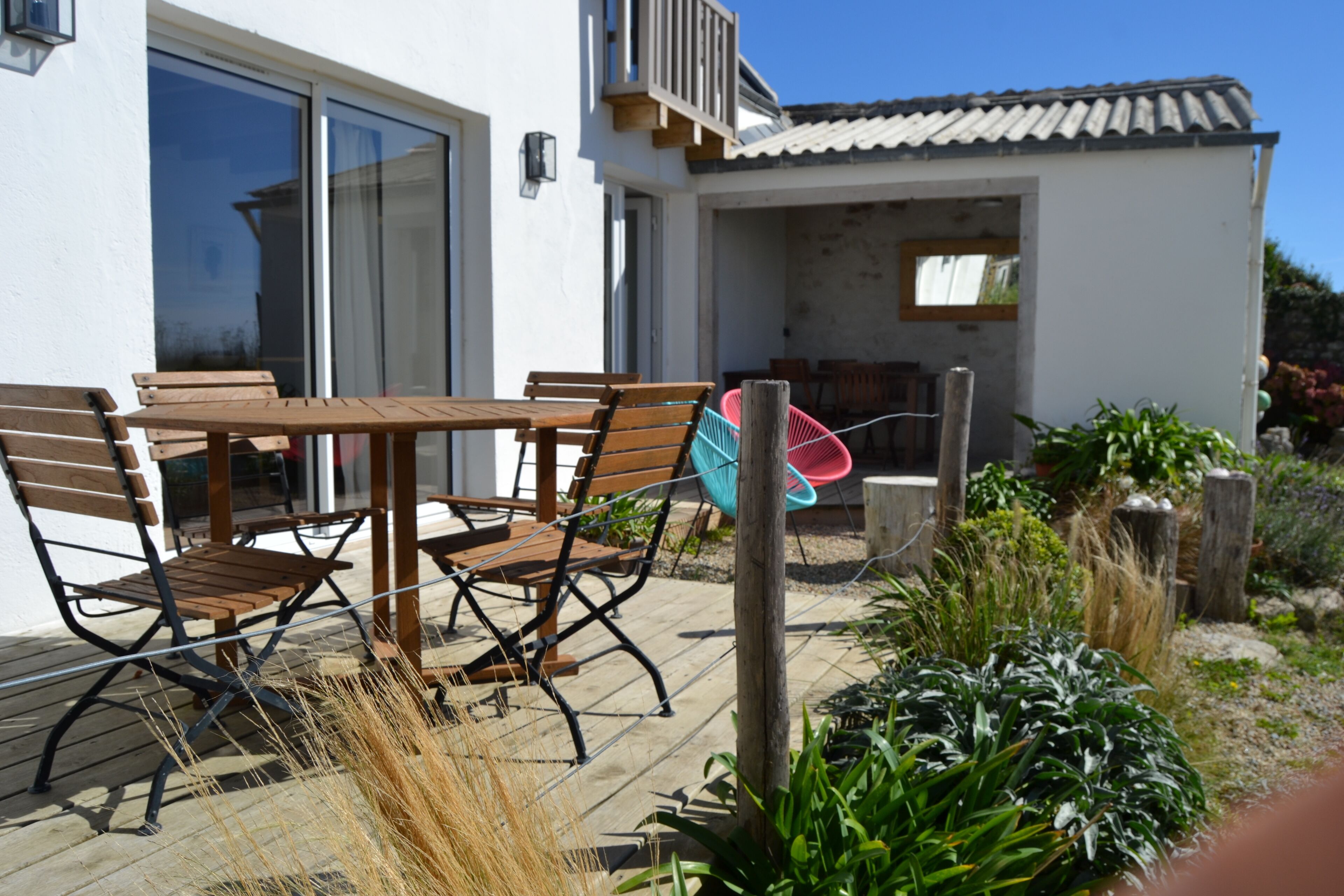 Une terrasse extérieure avec une table et des chaises en bois, un fauteuil suspendu et un petit jardin agrémenté de plantes et d’une clôture en bois.