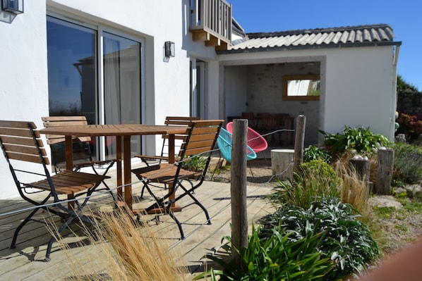Un patio exterior con una mesa y sillas de madera, una silla colgante y un pequeño jardín con plantas y una cerca de madera.