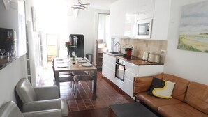 Une cuisine moderne avec des meubles blancs, un four à micro-ondes et un évier. On y trouve un coin repas avec une table dressée pour quatre personnes, ainsi qu’un canapé en cuir marron agrémenté d’un coussin jaune.