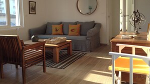 Un salon avec un canapé gris, une table en bois et une chaise. On y trouve une fenêtre, un miroir sur le mur et une porte qui donne sur une autre pièce.