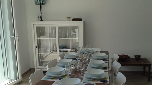 Une table à manger est dressée avec des assiettes, des verres et des couverts, entourée de chaises blanches dans une pièce bien éclairée. On aperçoit, au fond, un meuble et une lampe.