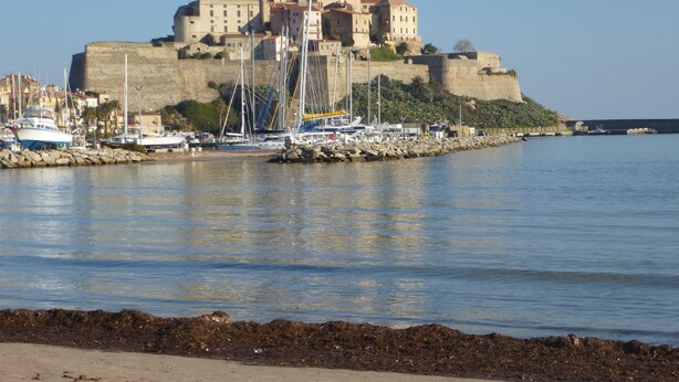 Un pueblo costero con una imponente estructura similar a un castillo, un puerto deportivo con varias embarcaciones y una playa arenosa en primer plano.