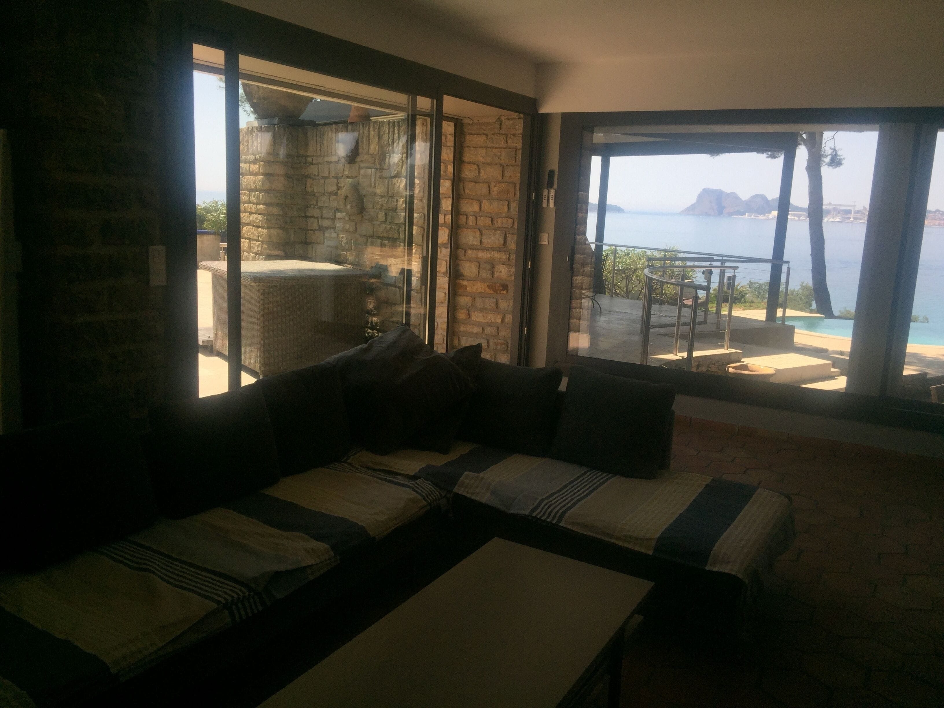 A room with a stone wall, a sofa with striped cushions, and a view of a balcony with a pool and mountains.