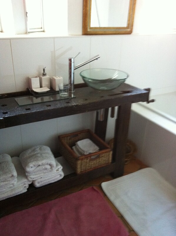 A bathroom with a glass bowl sink, a mirror, and a shelf with towels and toiletries.