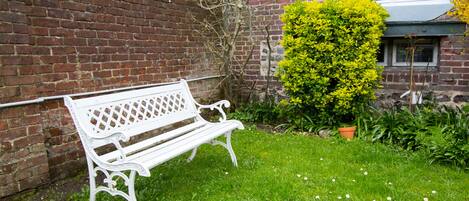A white bench in a grassy area with a brick wall and a hedge in the background.