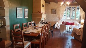 A dining area with a wooden table set for six, surrounded by wooden chairs. There is a fireplace with a mantel above the table. A sofa and a lamp are visible in the background, with a window showing greenery outside. - Country house, quiet location, private pool. Country house. Magnificent view. (Montcuq en Quercy Blanc)