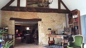 A rustic room with exposed wooden beams, a stone wall, a wooden table with a lamp, a green armchair, and a bookshelf.