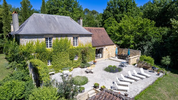 A stone house with a tiled roof, surrounded by lush greenery and a patio with outdoor seating.
