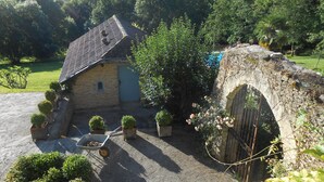 A stone house with a tiled roof, surrounded by lush greenery and a stone bridge.