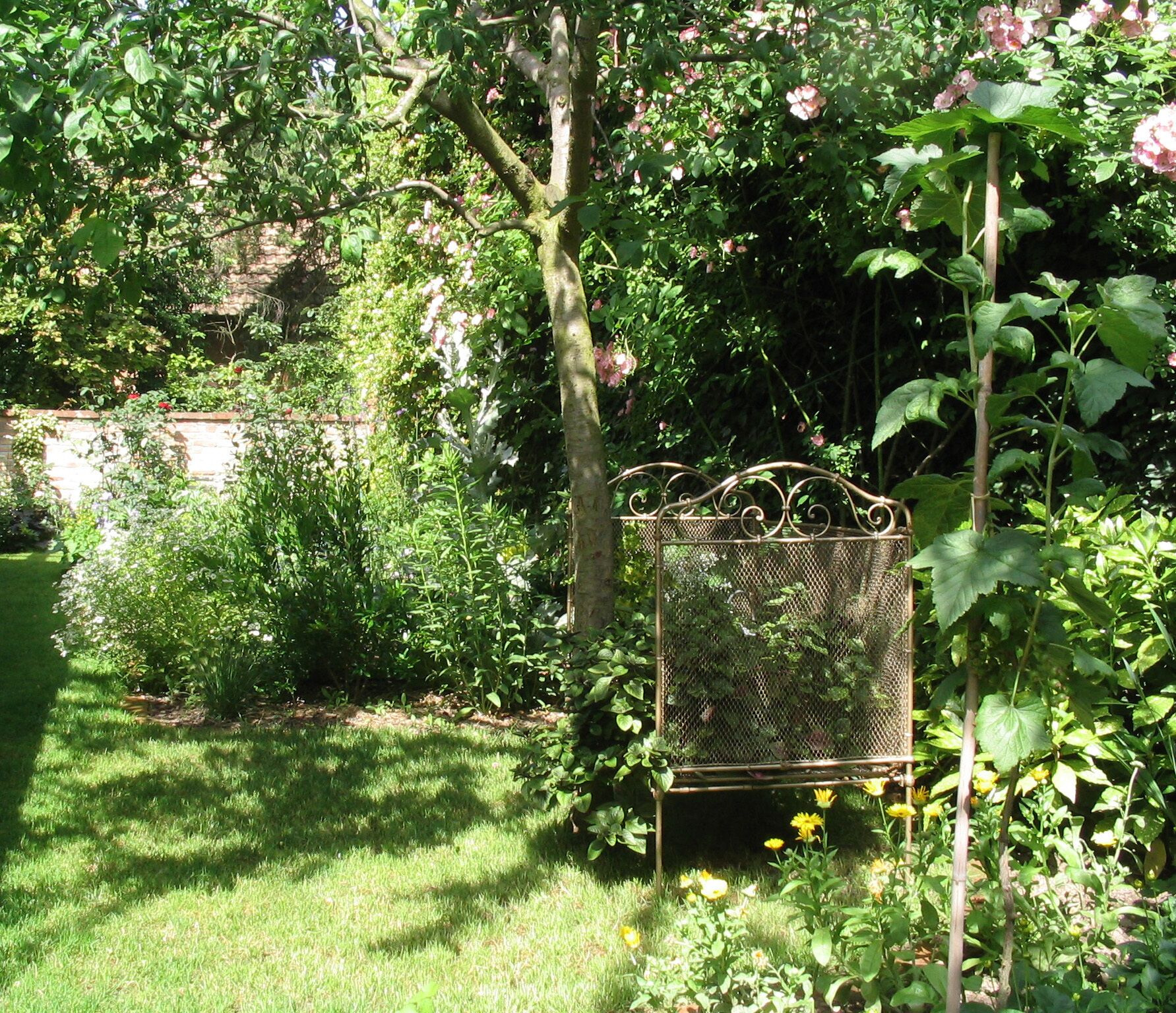 Een tuin met een metalen vogelvoerder, weelderig groen en een bakstenen muur op de achtergrond.