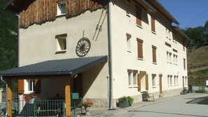 A two-story building with a sloped roof, a wooden upper section, and a beige lower section. There is a covered porch with a bench and potted plants. - Vaujany, alpes d'huez, ski, bike, hike, trail chalet 40m2 2/5p, (Vaujany)