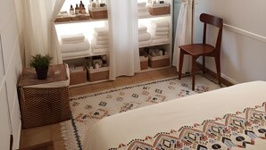 Un dormitorio con cama, una silla, estante con artículos de tocador y vistas a través de una ventana con cortinas.
