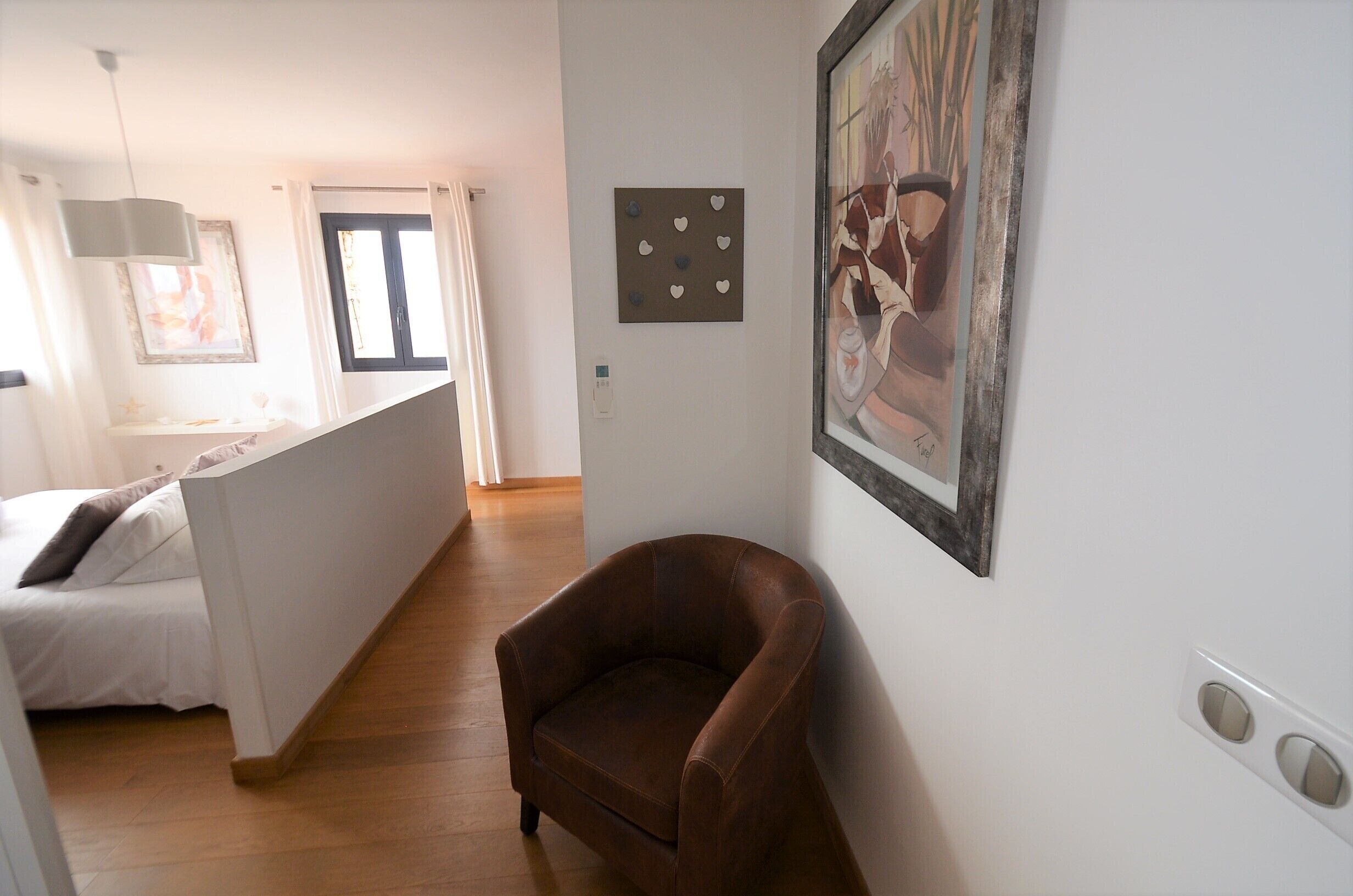Een slaapkamer met een bed, een bruine fauteuil, een schilderij en een raam met gordijnen.
