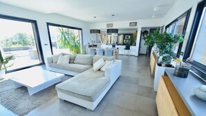 A modern living room with a sectional sofa, a coffee table, and a dining area with chairs. There is a kitchen in the background with a large island and modern appliances.