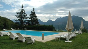 A swimming pool with lounge chairs and umbrellas, surrounded by greenery and mountains.