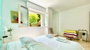 A bedroom with a large bed, a window with a view of greenery, a chair, a table with a striped tablecloth, and a mirror.
