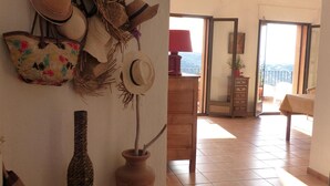Una habitación con suelo de baldosas, un banco de madera, un perchero de pared con sombreros y un bolso decorativo, una planta en maceta y un acceso a un balcón con vistas.