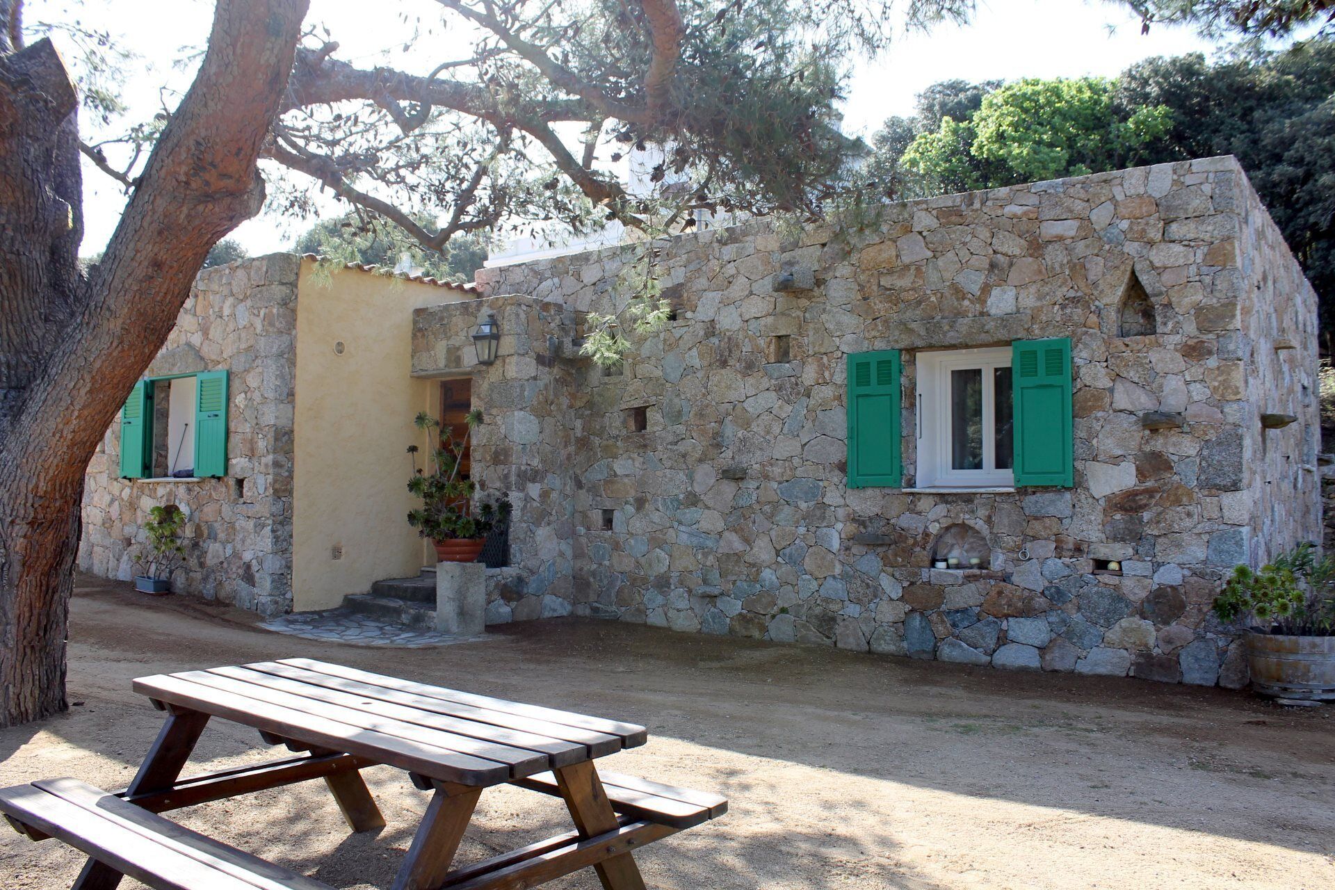 A stone building with green shutters, a wooden bench, and potted plants.