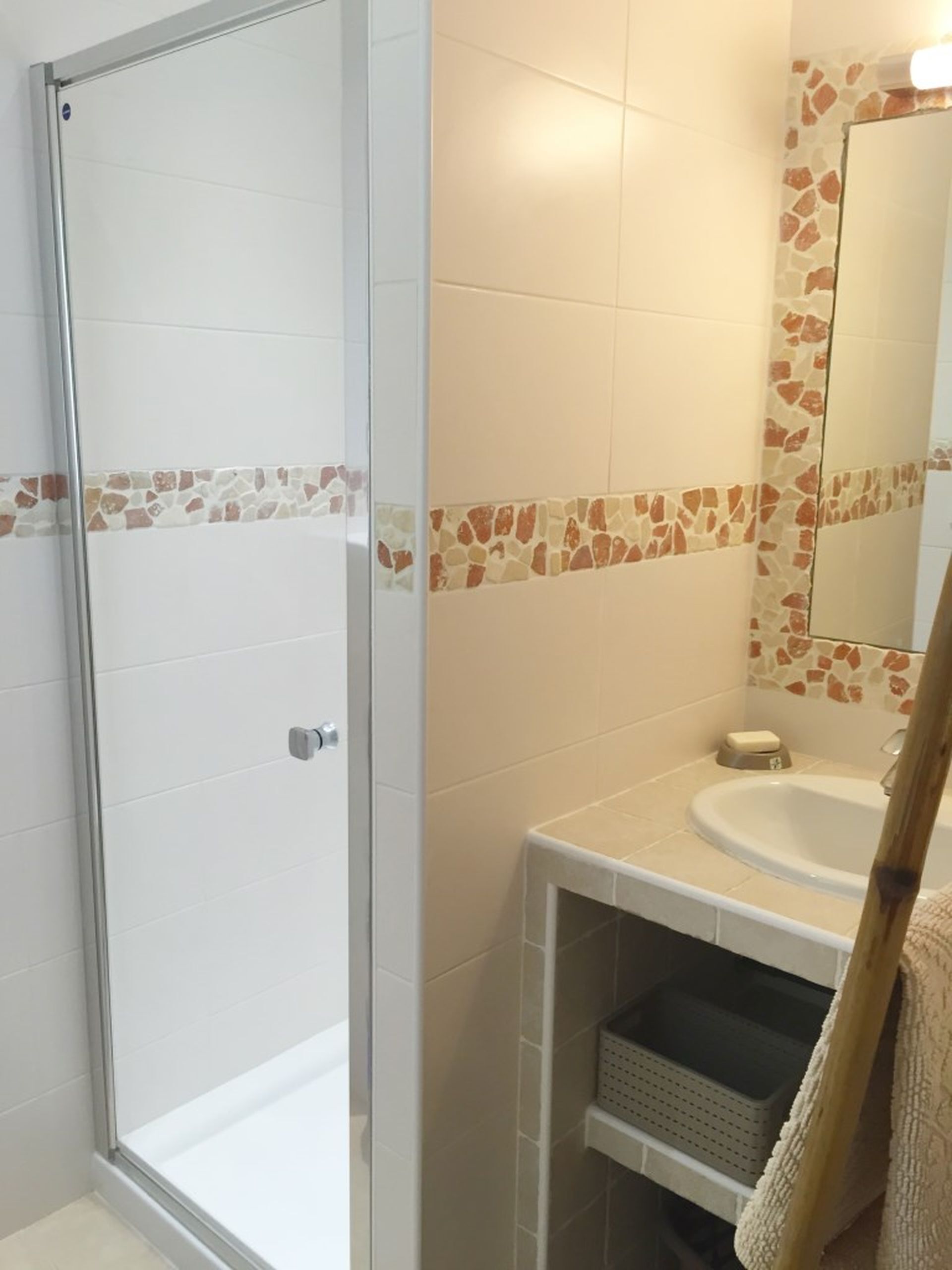 Een badkamer met een glazen douchedeur, een wastafel met spiegel en een betegelde muur met mozaïekrand.