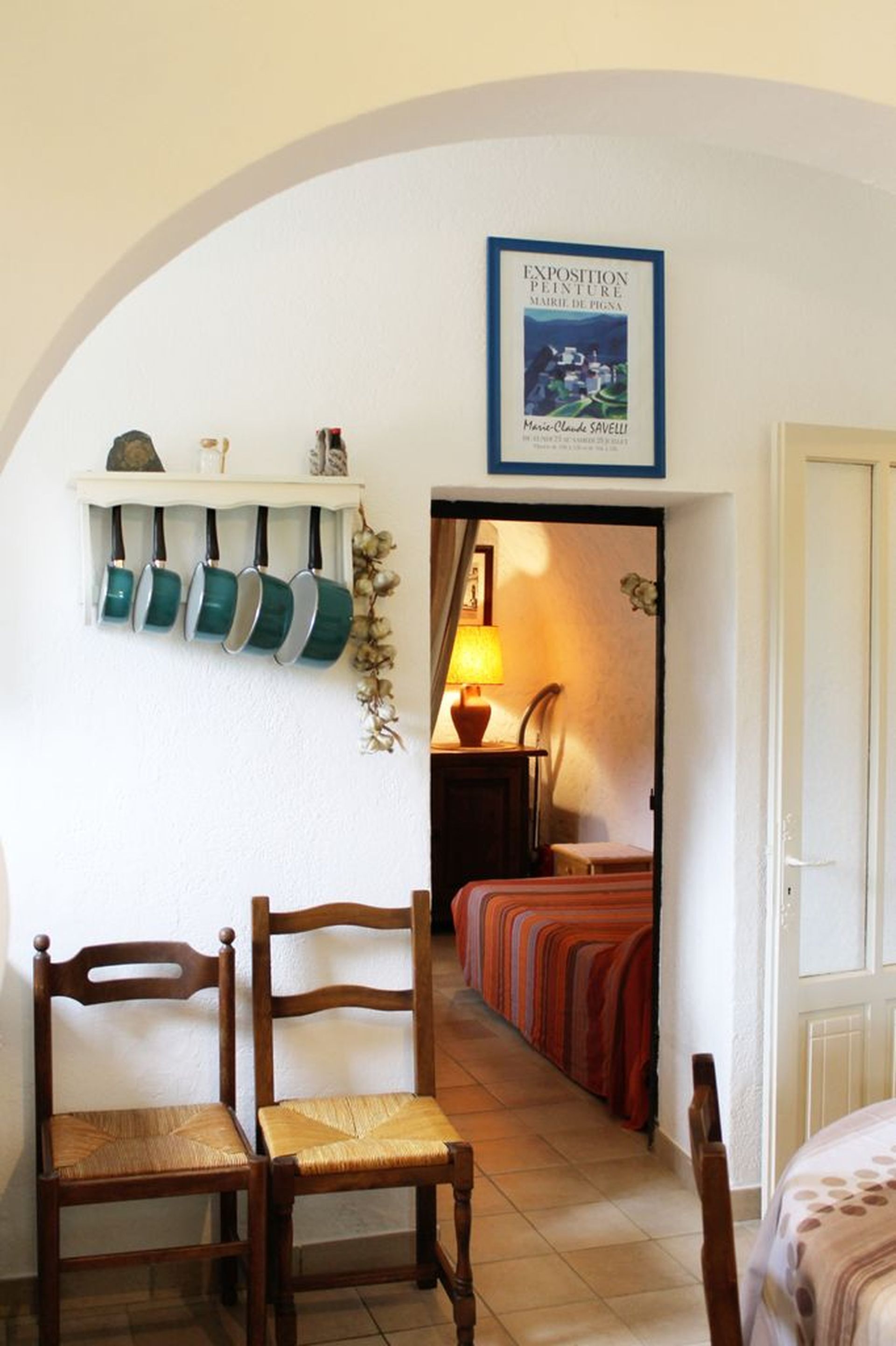 Una stanza con una sedia di legno, un letto con coperta a righe, una mensola con tazze verdi e un quadro appeso al muro.