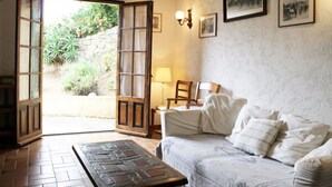Habitació acollidora amb llit, cadira i taula. Hi ha una porta que dona a una zona exterior amb plantes.