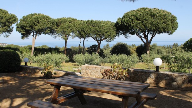Una taula de picnic de fusta amb bancs, envoltada de murs de pedra i diverses plantes, amb vista a un paisatge maritim idíl·lic.