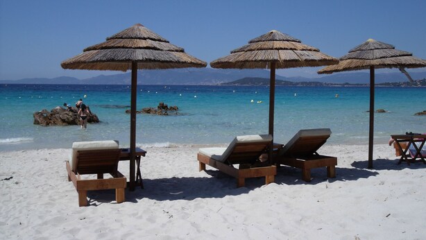 Una playa con sillones de madera y sombrillas de paja, un mar azul cristalino y afloramientos rocosos al fondo.