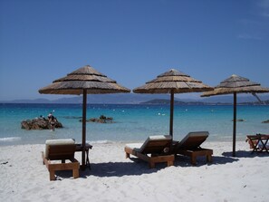 A beach with wooden lounge chairs and thatched umbrellas, a clear blue sea, and rocky outcrops in the background.