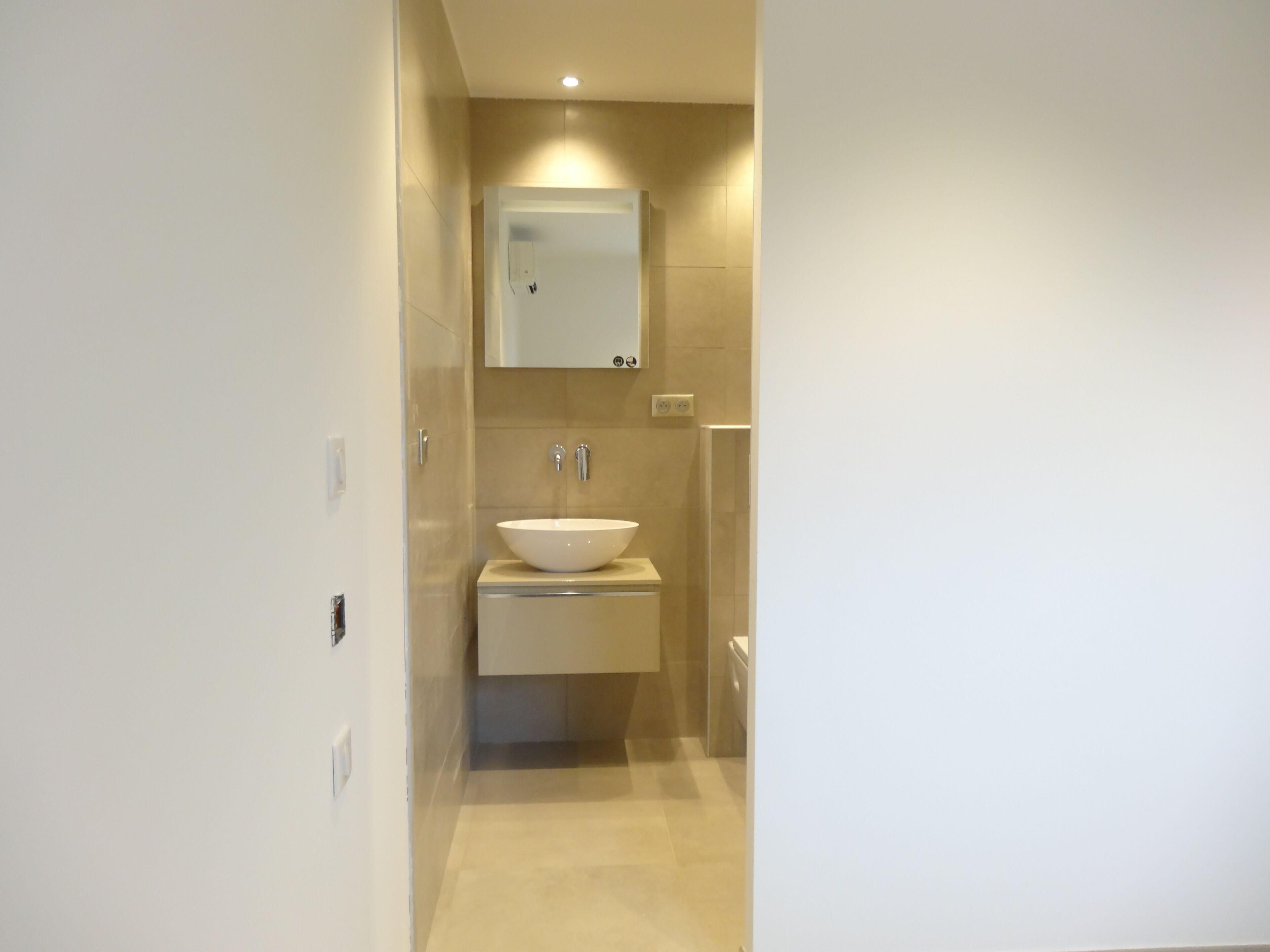 Een moderne badkamer met een witte wastafel, een spiegel erboven en beige tegels op de wanden en vloer.